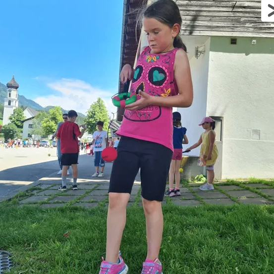 Ein junges Mädchen in einem rosa Tanktop steht auf Gras und hält einen roten Ball. Es ist von Kindern und Erwachsenen in der Nähe eines Gebäudes umgeben.