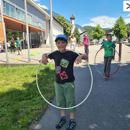 Ein kleiner Junge steht auf einem Bürgersteig und hält einen Hula-Hoop, während ein Junge hinter ihm einen größeren hält. Im Hintergrund befinden sich Gebäude und Bäume.