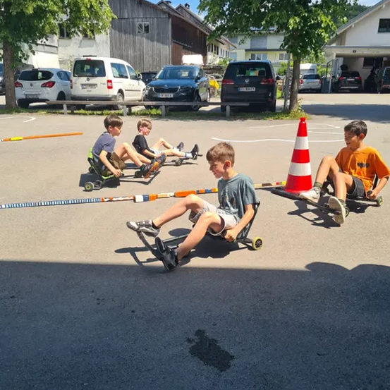 Vier Jungen fahren Go-Karts auf einem Parkplatz, zwei sitzen, während die anderen liegen. Ein orangefarbener Verkehrskegel steht in der Mitte. Hinter ihnen sind Autos und Häuser geparkt.