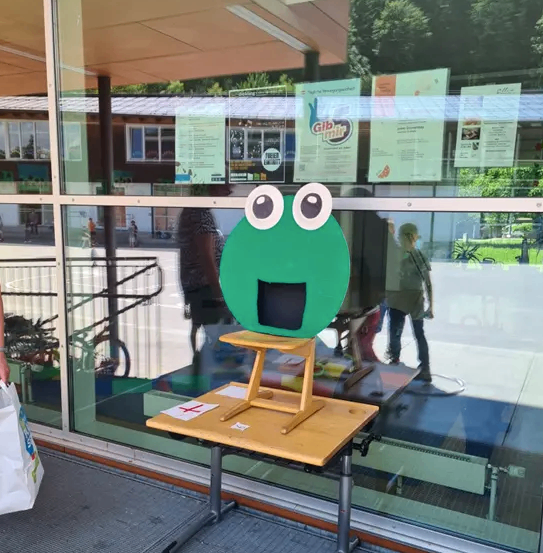 Ein grüner Frosch mit großen Augen steht auf einem Holztisch vor einer Glaswand mit Flyern. Leute gehen draußen vorbei.