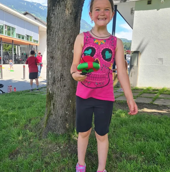 Ein Mädchen in einem rosa Shirt steht in einem Park, lächelt und hält ein grünes und rotes Spielzeug. Hinter ihr ist ein Gebäude mit Fahrrädern und Menschen zu sehen.