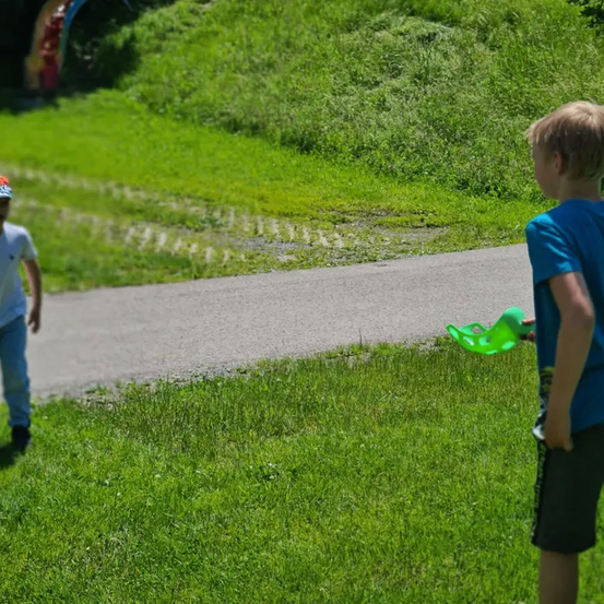 Zwei Jungen stehen auf einem Grasweg. Einer hält ein grünes Objekt, vermutlich ein Frisbee. Der Junge auf der linken Seite trägt einen Hut und Turnschuhe. Hinter ihnen ist ein Grashügel zu sehen.