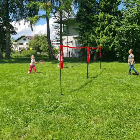 Zwei Kinder spielen an einem sonnigen Tag Badminton in einem grünen Park mit einem Haus im Hintergrund.