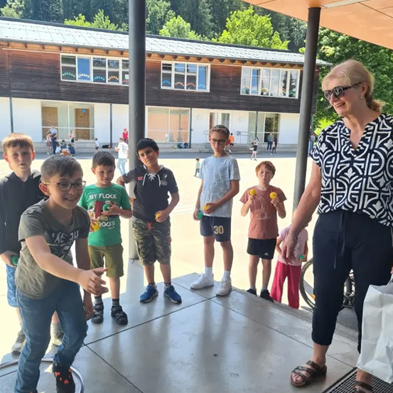 Eine Gruppe von Kindern und eine erwachsene Frau stehen unter einem Schutzdach auf einem Schulhof. Die Kinder halten kleine Gegenstände, möglicherweise Bälle, und scheinen an einem Spiel teilzunehmen. Die Frau lächelt und schaut die Kinder an.
