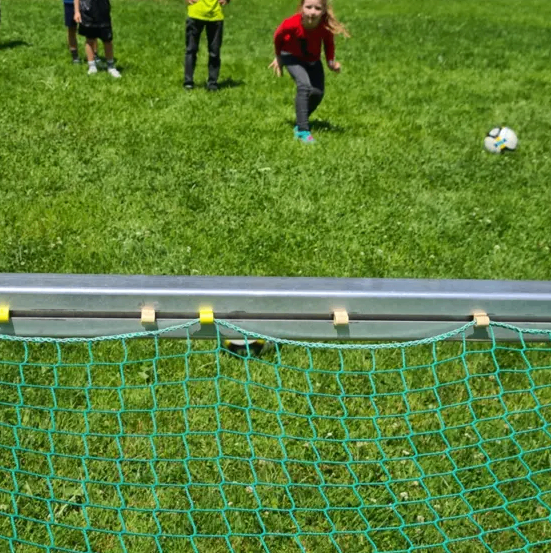 Ein Mädchen in einem roten Shirt läuft auf einen Fußball auf einem Rasenfeld zu, mit einem Jungen in einem gelben Shirt im Hintergrund.