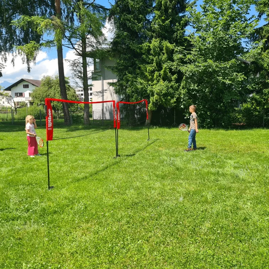 Zwei Kinder spielen Badminton auf einem Rasenfeld mit einem roten Netz. Sie halten Schläger und sind von Bäumen und einem Gebäude umgeben.