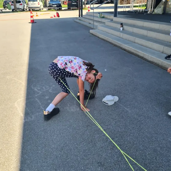Ein Mädchen beugt sich vor, hält eine Schnur fest, mit einer weißen Kappe in der Nähe, auf einem Parkplatz mit Gebäuden und Bäumen im Hintergrund.