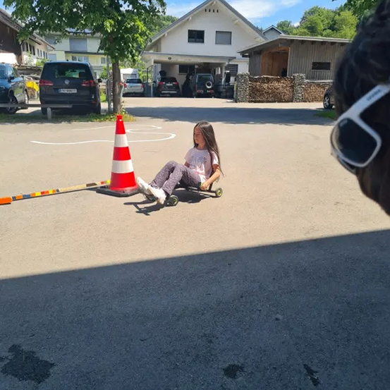 Ein kleines Mädchen fährt auf einem Skateboard und navigiert eine Kurve, die durch einen Kegel und eine Linie markiert ist. Hinter ihr sind Häuser und Autos zu sehen.