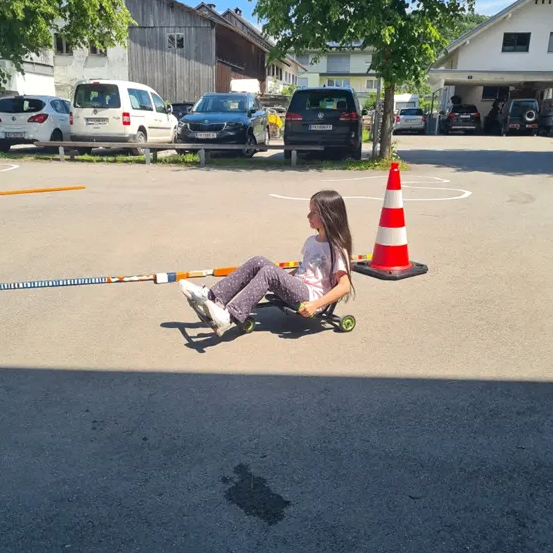 Ein Mädchen fährt mit einem Go-Kart auf einer asphaltierten Straße, in der Nähe eines Verkehrskegels. Hinter ihr sind mehrere Autos geparkt, und ein Gebäude ist im Hintergrund.
