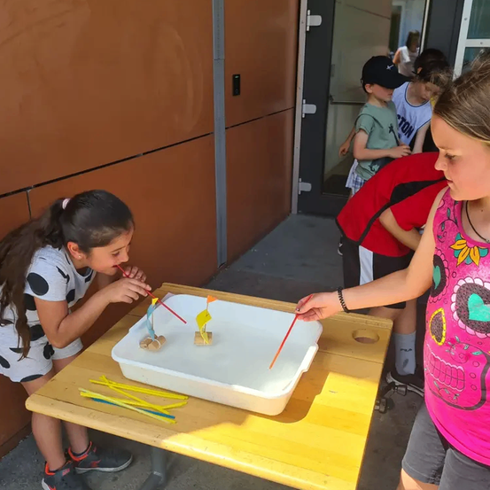 Zwei Mädchen pusten auf Strohhalme, um Boote auf einem Tisch zu bewegen, während andere Kinder zusehen. Die Boote sind klein und aus Holz.