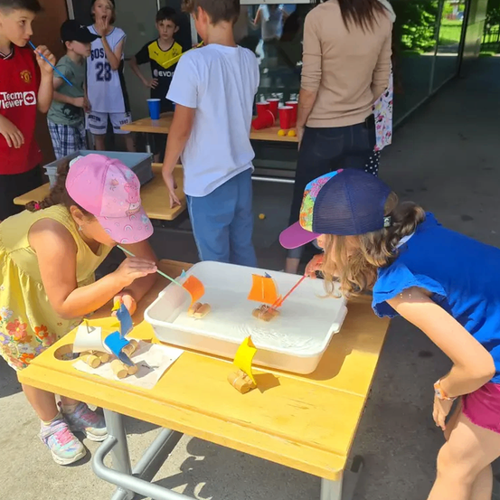 Mehrere Kinder beschäftigen sich mit einer Aktivität mit Booten auf einem Tisch in einem Raum, in dem andere Kinder und Erwachsene im Hintergrund sind.