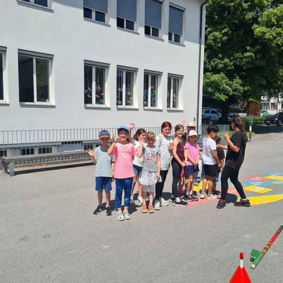 Eine Gruppe von Kindern und Erwachsenen steht vor einem weißen Gebäude mit Fenstern und einem Zaun. Die Kinder tragen Freizeitkleidung und einige tragen Hüte. Sie posieren für ein Foto.