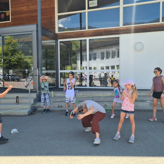 Eine Gruppe von Kindern und Erwachsenen spielt draußen vor einem modernen Gebäude mit Glaswänden. Die Kinder halten Seile und einer beugt sich vor.