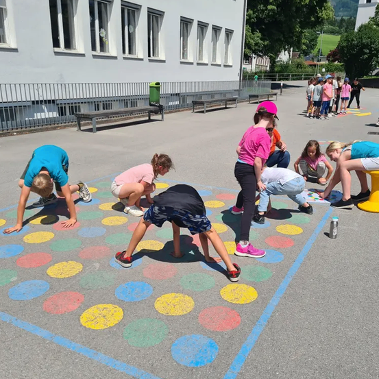 Kinder spielen Hüpfspiel auf einem Schulhof. Sie tragen Freizeitkleidung. Einige tragen Turnschuhe, während andere barfuß sind. Sie sind draußen in der Nähe eines Gebäudes.