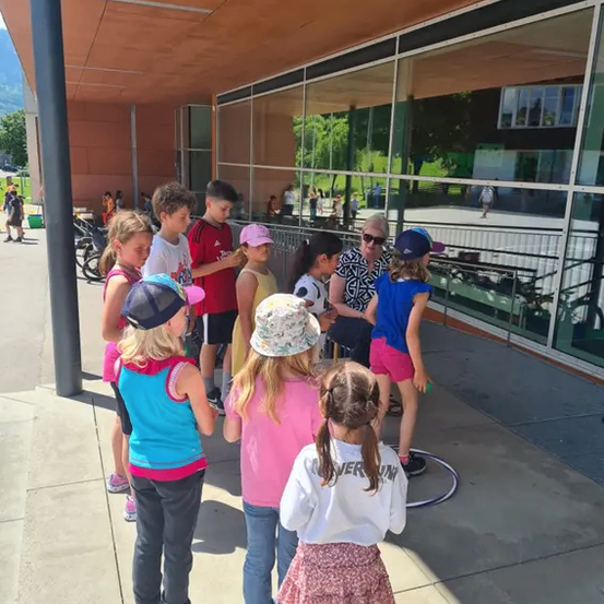 Eine Gruppe von Kindern und einer Frau steht vor einem Gebäude mit Glaswänden, Fahrrädern und Bäumen im Hintergrund.