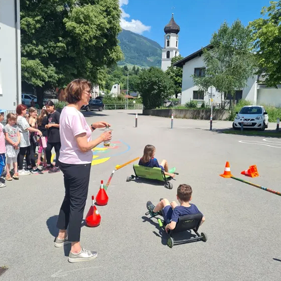 Eine Frau steht auf einem Parkplatz mit Kindern. Zwei Kinder sitzen in Go-Karts, während sie von Verkehrskegeln umgeben sind. Gebäude und Bäume sind im Hintergrund.