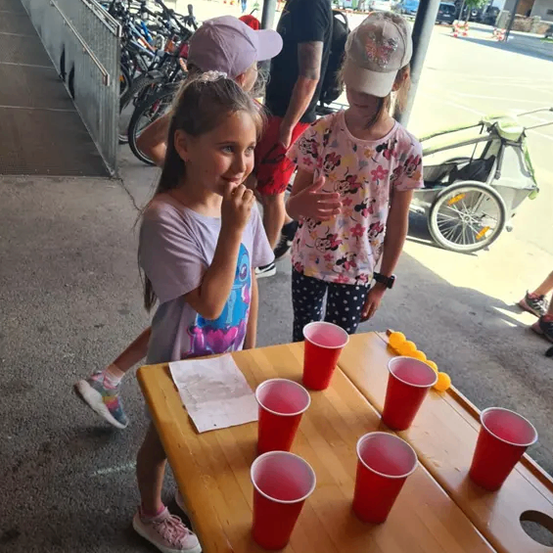Zwei Mädchen in Mützen spielen ein Bierpong-Spiel mit roten Bechern auf einem Holztisch, während ein Mann hinter ihnen steht.