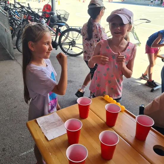 Zwei Mädchen spielen Beer Pong auf einem Holztisch mit roten Bechern und gelben Bällen, in der Nähe eines Fahrradständers und eines Metallzauns.