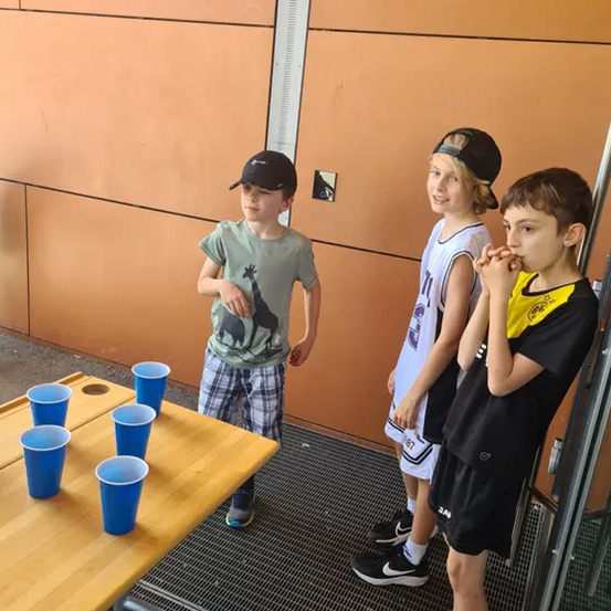 Drei Jungen stehen in der Nähe eines Tisches mit blauen Bechern und spielen möglicherweise ein Spiel. Ein Junge trägt einen Hut und Turnschuhe, ein anderer ein Basketballtrikot und der dritte ein T-Shirt mit Giraffenaufdruck.