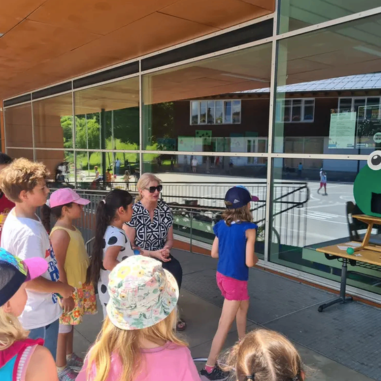 Eine Gruppe von Kindern steht vor einem Gebäude mit Glaswänden, eine Frau mit Brille und einer schwarz-weißen Bluse sitzt auf einer Bank und spricht mit ihnen.