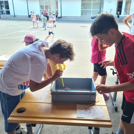 Zwei Jungen arbeiten an einem Projekt auf einem Tisch, während ein Mädchen mit einer rosa Mütze zusieht. Hinter ihnen spielen Kinder auf dem Spielplatz. Ein Gebäude mit Glaswänden befindet sich im Hintergrund.