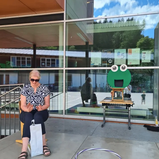 Eine Frau sitzt auf einem Stuhl vor einem Gebäude und schaut auf einen Tisch mit einem grünen Frosch-Ausschnitt. Hinter ihr spiegelt sich die Umgebung in einer Glaswand.