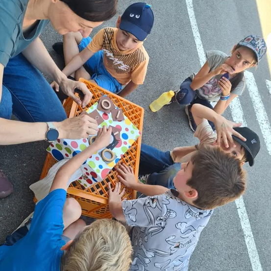 Eine Frau und mehrere Kinder sitzen in einem Kreis um einen Kisten mit Spielzeug. Ein Kind hält ein Spielzeug und schaut in die Kamera. Ein anderes Kind isst etwas. Die Frau hält eine Schachtel mit Spielzeug.