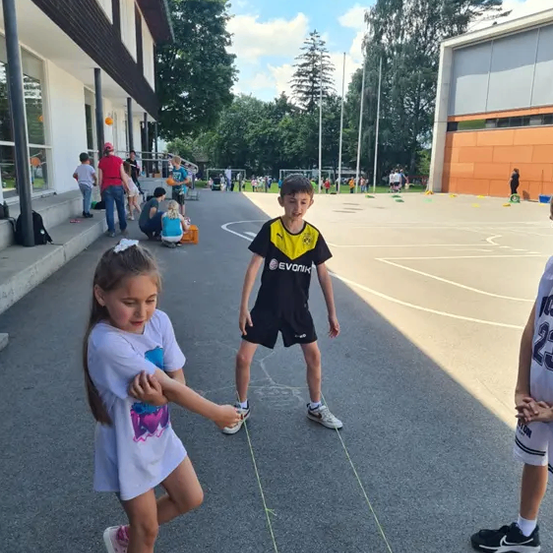 Ein junges Mädchen und ein Junge spielen ein Spiel auf einer Betonfläche und halten eine Schnur zwischen ihren Händen. Der Junge trägt ein schwarzes Trikot. Es gibt Leute, die auf dem Gras sitzen und andere stehen in der Nähe des Gebäudes.