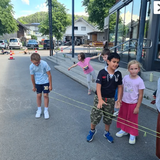 Fünf Kinder spielen draußen mit gelben Schnüren. Ein Junge hält eine Schnur, während ein anderer Junge herumrennt. Ein Mädchen steht vor einem Gebäude.
