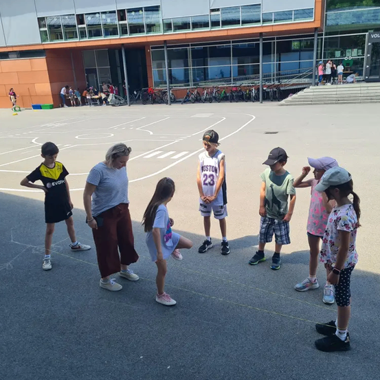 Eine Gruppe von Kindern und Erwachsenen steht auf einem Basketballplatz, wahrscheinlich beschäftigt mit einem Spiel oder einer Aktivität. In der Nähe sind Fahrräder geparkt.
