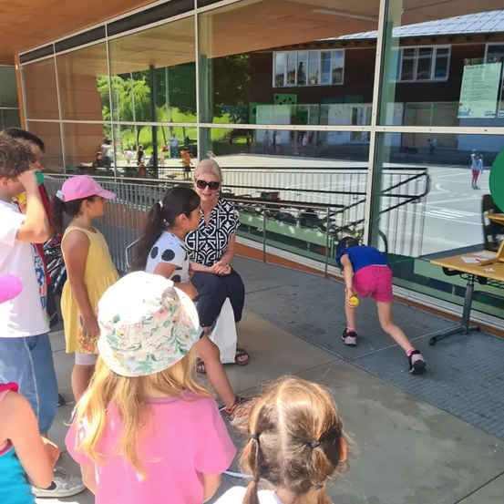 Eine Gruppe von Kindern und einer Frau versammelt sich vor einem Gebäude mit Glaswänden, eine Frau sitzt auf einer Bank und ein Mädchen spielt mit einem Ball.