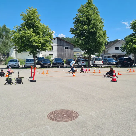 Mehrere Kinder nehmen an einem Hindernisparcours auf einem gepflasterten Platz teil. Bäume und geparkte Autos umgeben das Gelände. Ein Kind läuft auf die Kegel zu, während andere in der Nähe sitzen.