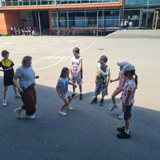Mehrere Kinder versammeln sich auf einem Schulhof, einige gehen und andere stehen. Im Hintergrund sind Fahrräder geparkt.