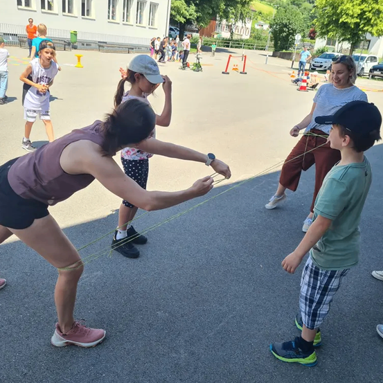 Eine Gruppe von Menschen ist im Freien versammelt. Einige spielen mit Seilen, während andere zusehen. Im Hintergrund befinden sich Bäume und ein Gebäude.