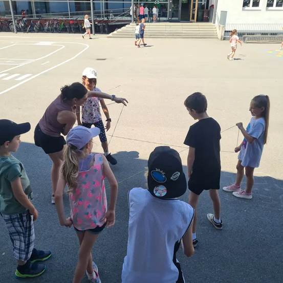 Eine Gruppe von Kindern und Erwachsenen ist draußen in einem gepflasterten Bereich. Einige halten Seile. Eine Frau unterrichtet sie. Es gibt Menschen, die in der Ferne gehen. Es sind Fahrräder am Rand geparkt.