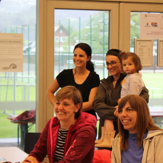Fünf Frauen stehen in einem Raum mit Glastüren und lächeln für ein Foto. Ein Kind wird von einer Frau mit Brille gehalten. Hinter ihnen hängen Poster an der Wand.