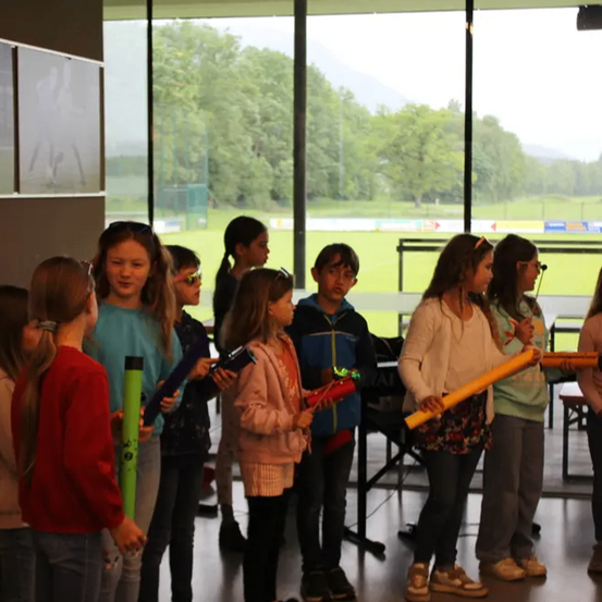 Eine Gruppe junger Mädchen in einem Klassenzimmer, die Instrumente, möglicherweise Flöten, halten, wobei ein Rasenfeld durch große Fenster sichtbar ist.