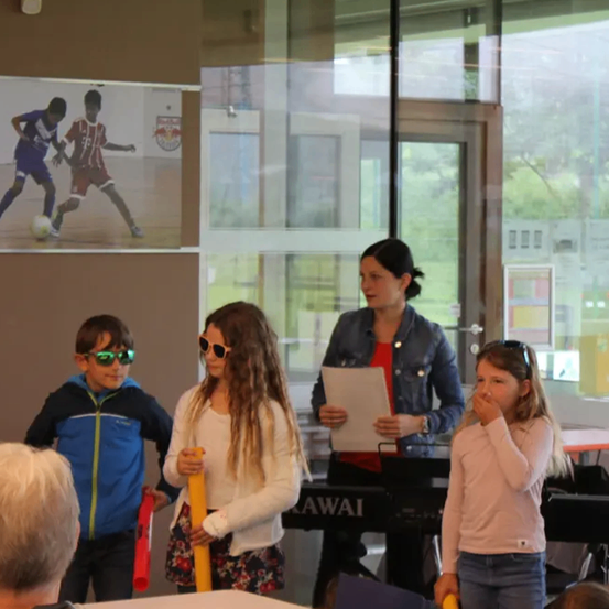 Eine Gruppe von Kindern und einer erwachsenen Frau stehen in einem Raum mit einem Fußballposter an der Wand, die Frau hält ein Papier und ein Kind hält einen roten Schläger.