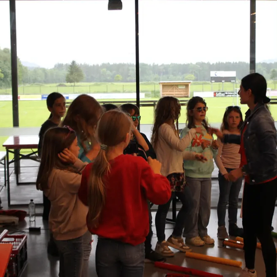 Eine Gruppe von Mädchen in verschieden farbigen Oberteilen und Hosen steht in einem glasverkleideten Raum. Sie scheinen einer Lehrerin zuzuhören, die eine Jeansjacke und Turnschuhe trägt. Ein Tisch mit einer Flasche und einem Korb befindet sich in der Nähe.