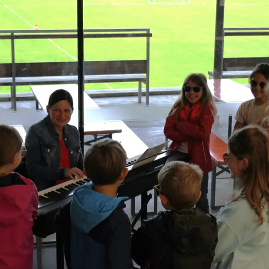 Eine Frau spielt ein Keyboard, während Kinder zusehen, mit Blick auf ein Stadion im Hintergrund. Die Kinder tragen Brillen und Jacken, und die Frau lächelt.