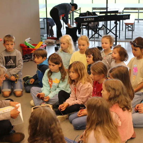 Eine Gruppe von Kindern sitzt im Kreis und hört einer erwachsenen Frau zu, die ein Buch in der Hand hält. Im Hintergrund ordnet ein Mann ein Keyboard an.