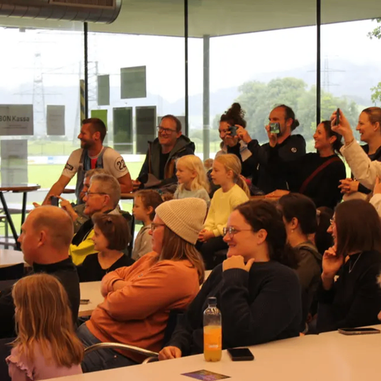 Eine Gruppe von Menschen sitzt an Tischen in einem glasumschlossenen Raum, mehrere machen Fotos. Eine Frau hält eine Flasche Saft. Ein Schild steht für BON Kassa.