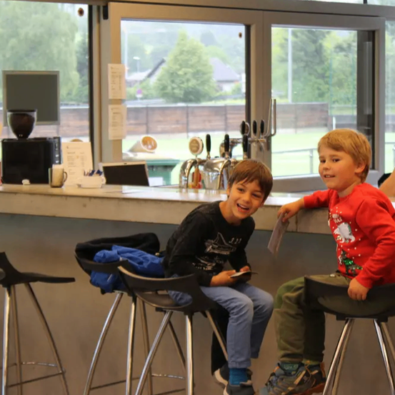 Zwei Jungen sitzen auf Barhockern, einer in einem roten Pullover, der andere in Schwarz. Ein Mann steht hinter ihnen. Eine Kaffeemaschine, Papiere und Tassen sind auf der Theke. Fenster zeigen ein grünes Feld draußen.