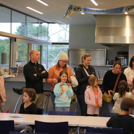 Eine Gruppe von Erwachsenen und Kindern steht in einem Raum mit einem Banner, auf dem ERNE FC SCHLINS steht. Sie schauen in die gleiche Richtung. Eine Frau vorne hält ein rosa Tuch. Eine andere Frau hält eine Tasche. Der Raum hat einen Küchenbereich mit einer Mikrowelle und einer Glaswand.