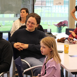 Eine Frau mit Brille sitzt mit einem Kind und lächelt in die Kamera. Sie sind an einem Tisch mit anderen Erwachsenen, nahe einer Glaswand.