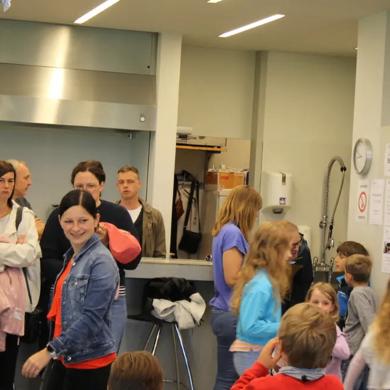 Eine Gruppe von Menschen steht in einem Flur. Eine lächelnde Frau in einer Jeansjacke steht vorne. Hinter ihr schauen mehrere andere in verschiedene Richtungen. Eine Uhr und einige Aushänge sind an der Wand.