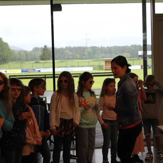 Eine Gruppe von Kindern und einer Frau steht drinnen, mit einem landschaftlichen Außenblick. Die Kinder tragen Sonnenbrillen, und die Frau hält eine Kamera. Der Außenbereich zeigt ein grünes Feld, Bäume und ein Gebäude.