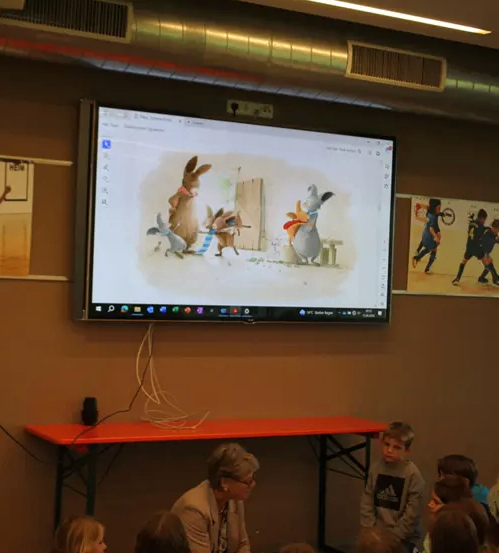 Eine Gruppe von Kindern sitzt um eine Frau vor einem Tisch und schaut auf einen an der Wand montierten Monitor, der eine animierte Szene zeigt.