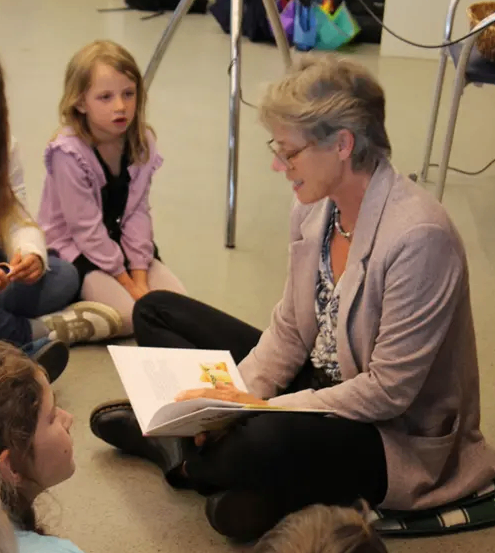 Eine Frau liest Kindern ein Buch vor. Sie sitzt auf dem Boden. Die Kinder sitzen im Kreis um sie herum. Ein Kind schaut sie an, während ein anderes etwas anderes ansieht. Die Frau trägt eine Brille und eine Halskette. Hinter ihnen stehen Taschen und ein Stativ.