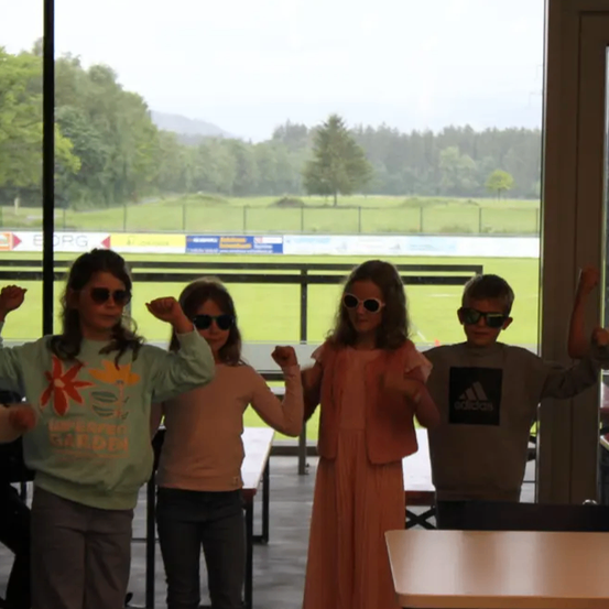 Fünf Kinder sind drinnen und tragen Sonnenbrillen. Sie gestikulieren mit den Händen. Dahinter ist ein Blick auf ein Grasfeld und Bäume zu sehen.